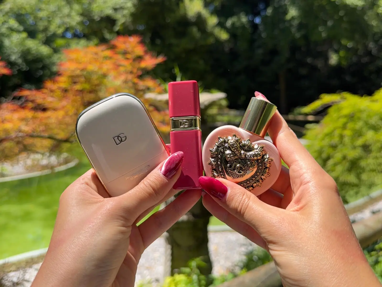 A creator’s hands holding a Dolce & Gabbana Beauty compact powder, lipstick and heart-shaped perfume in a garden