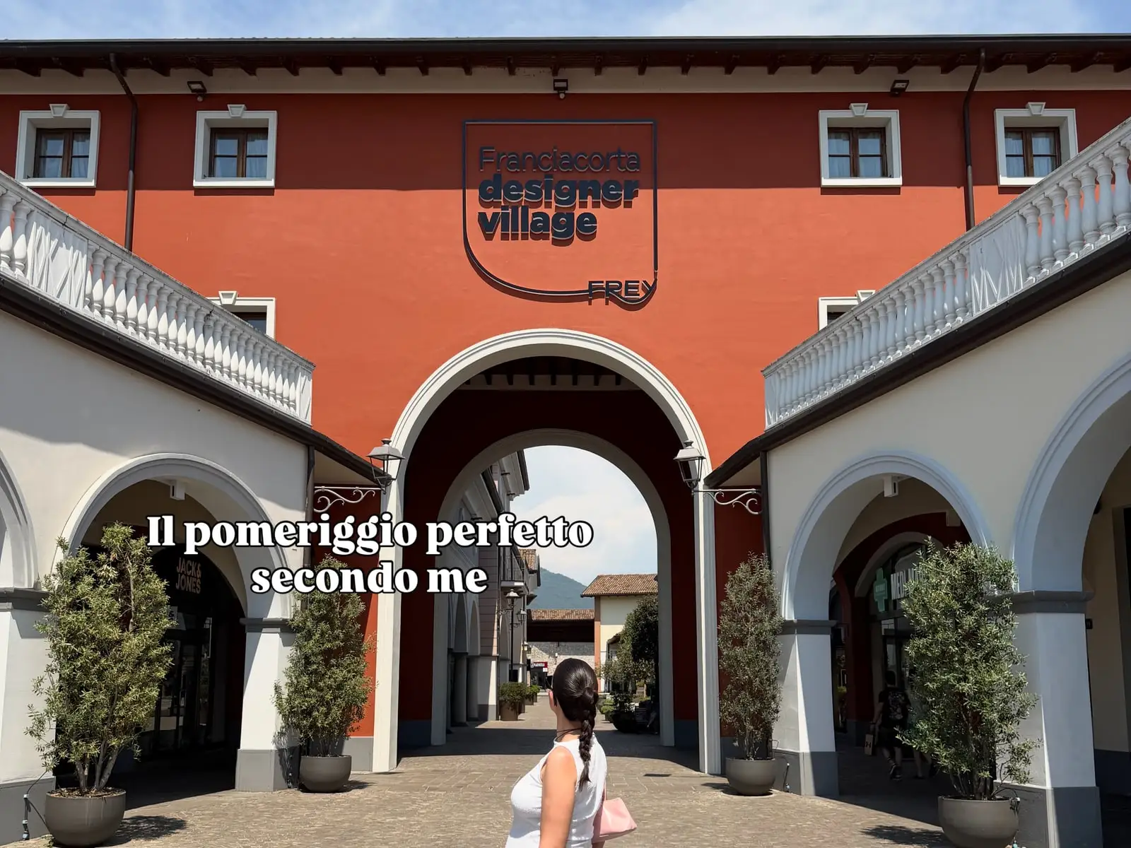 Entrée en arche de Franciacorta Designer Village avec son enseigne, l’inscription « Il pomeriggio perfetto secondo me » et une créatrice de dos