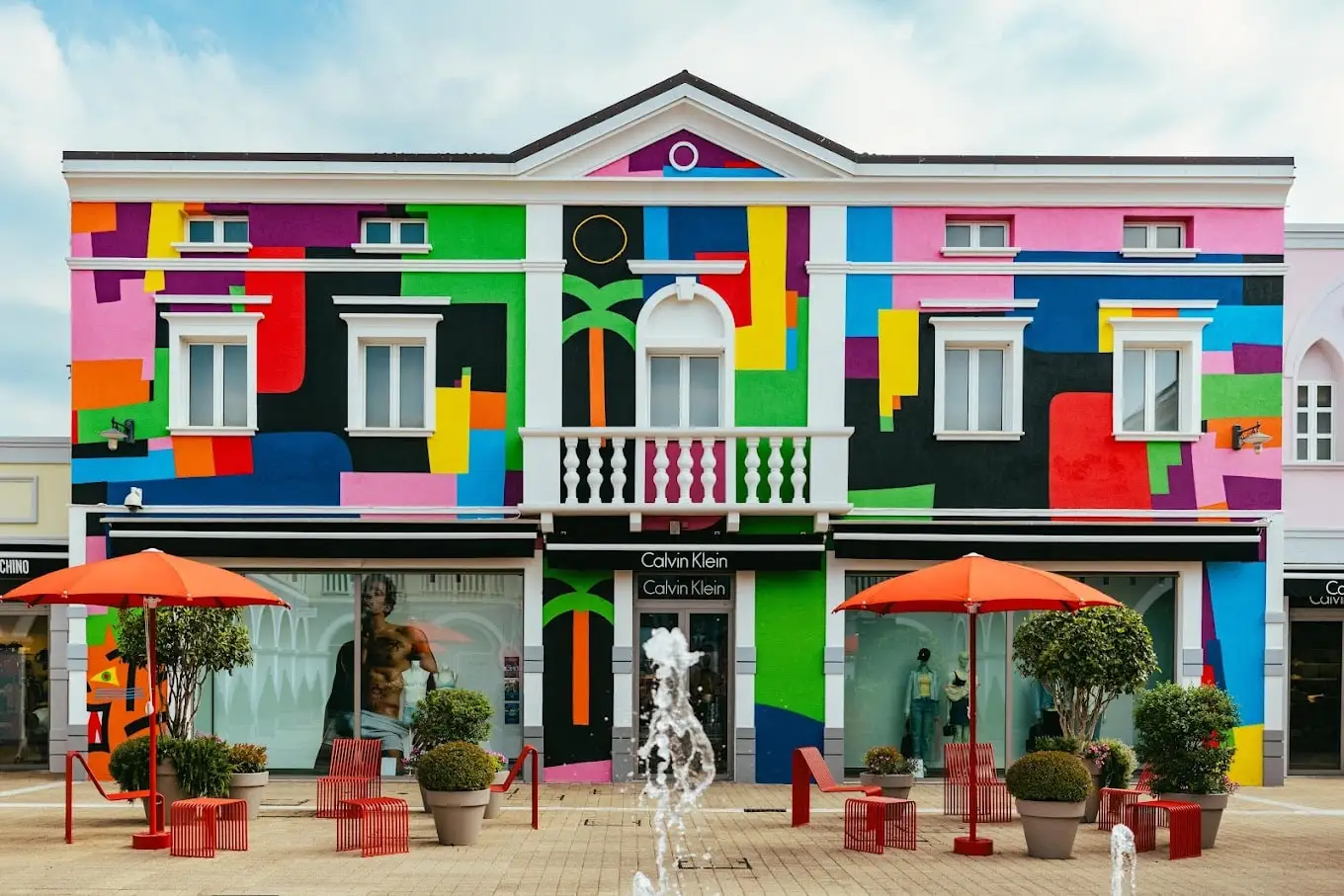 Façade de Palmanova Designer Village peinte en blocs de couleur, avec des parasols orange et une fontaine