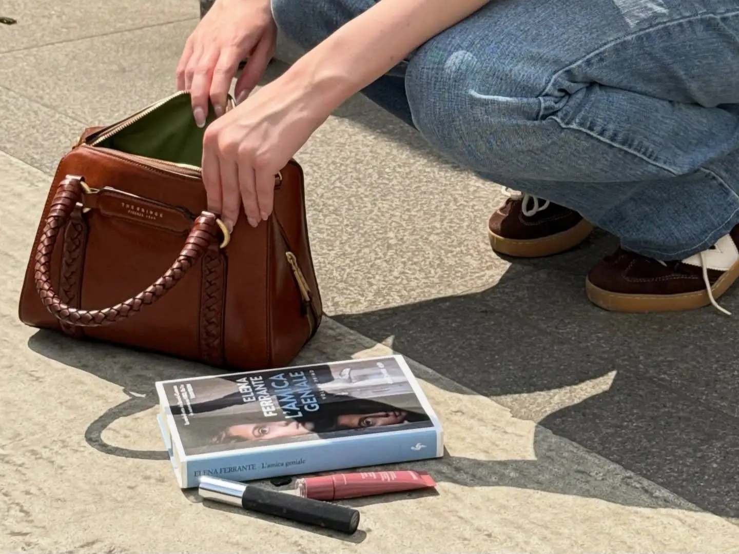 Creator crouching on a pavement opening a tan leather bag, next to a book and a lipstick